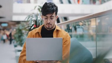 Yoğunlaşmış genç Arap adam dizüstü bilgisayarla oturmuş e-posta kontrol ediyor sosyal ağ sayfasına bakıyor bilgisayar uygulaması sohbet mesafesi kullanıyor çevrimiçi dükkânda kameraya bakıp gülümsüyor pozu veriyor.