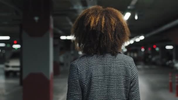 View from behind beautiful young girl business woman lady african american female with curly hair stylish hairstyle in gray coat walks in parking lot turns around looking at camera leing friendly — Stockvideo