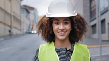 Kadın portre işçisi mesleği kıvırcık saçlı, inşaat mühendisi profesyonel Afro-Amerikan kadın şehir sokaklarında güvenlik kaskı takıyor ve gülümseyen dostça gülüşlere bakıyor.