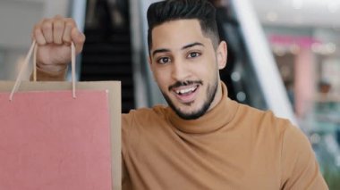 Alışveriş merkezinde gülümseyen genç Arap adam elinde alışveriş torbalarıyla elinde hediye paketlerine işaret ederek indirimli satışlara seviniyor. Tamam işaretleri onaylama jesti başarılı.