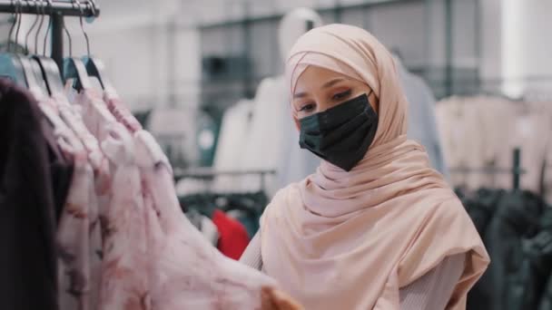 Jovem árabe mulher cliente comprador no hijab vestindo máscara médica protetora na loja de roupas escolhe cuidadosamente roupas em busca de roupa adequada olhando para o material de qualidade de compras à venda com desconto — Vídeo de Stock