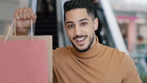 Feliz joven árabe hombre comprador de pie en el centro comercial sonriendo sosteniendo bolsas de compras en la mano señalando dedo índice en paquetes de regalo regocijo en descuentos venta mostrando ok signo de aprobación gesto de compra exitosa — Vídeos de Stock