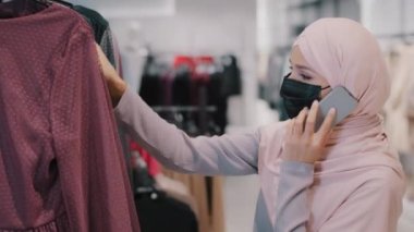 Tıbbi maske takan genç Arap kadın, elbise mağazasında elbise seçiyor. Telefonla yeni bir görünüm seçiyor. Satılık giysiler alıyor.