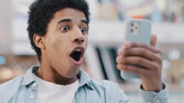 Afro-amerikan şaşkın erkek kullanıcı şaşırdı. Cep telefonuna bakan genç adam sürpriz bir sevinçle ağzını açtı. Zafer indirimi teklifi akıllı telefon mesajlarında şok edici haberler okuyor.
