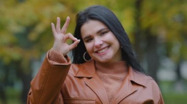 Portre mutlu genç İspanyol kadın model dışarıda duruyor gülümseyen beyaz dişli gülümseme kameraya bakıyor tamam işaret desteği onayını gösteriyor iyi bir takdir sembolü.