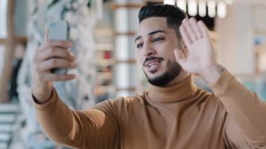 Genç adam blogcu elinde akıllı telefonuyla evde duruyor. Web kamerasından konuşuyor. Video görüşmeleri yapıyor. Telefondaki mobil uygulamayı kullanıyor.