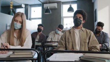 Tıp maskeli genç Afrikalı Amerikalı öğrenci, çok ırklı sınıf arkadaşlarının arasında masada oturuyor ve karantinadaki kısıtlamalarla birlikte grup semineri sırasında öğretmenlere soru soruyor.