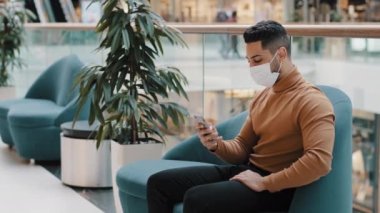 Tıbbi maskeli genç adam kapalı bir yerde oturuyor ve akıllı telefonuyla sosyal ağda mesajlar yazıyor. İnternet sitesinde internet sitelerinde sörf yapmak için mobil uygulama kullanarak e-posta gönderiyor.