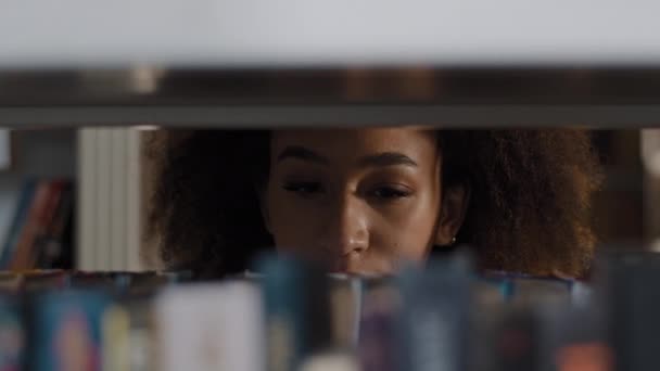 Joven estudiante afroamericana niña buscando cuidadosamente un libro adecuado en la estantería que se concentra en elegir el libro de texto adecuado para el estudio o el trabajo búsqueda de tareas literatura favorita en la biblioteca universitaria — Vídeos de Stock