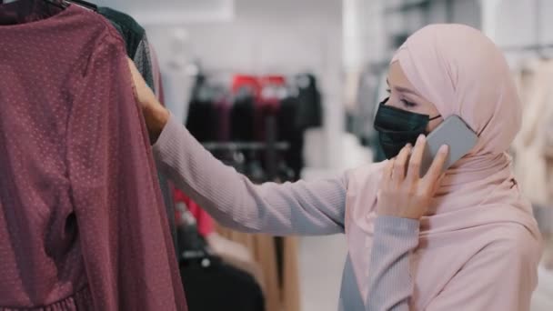 Young arab woman in hijab wearing medical mask chooses dress in clothing store speaks on phone consults about choosing new look buys clothes on sale communicates answers mobile call using smartphone — Stock Video