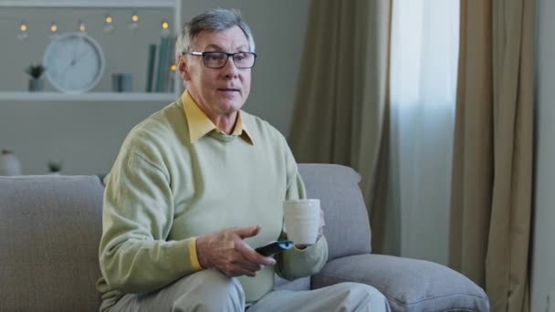 Homme âgé aux cheveux gris dans des lunettes assis sur le canapé regarder la télévision profiter de détente à la maison boire du thé vieux café mâle utilise la télécommande pour changer de chaînes souriant profiter drôle programme de télévision de film — Video