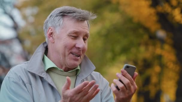 Радісний літній чоловік, що робить відеодзвінок, використовує сучасний смартфон, зрілий усміхнений дідусь, що тримає мобільний телефон, весело розмовляє, ділиться добрими новинами з друзями в концепції віддаленого зв'язку осіннього парку — стокове відео