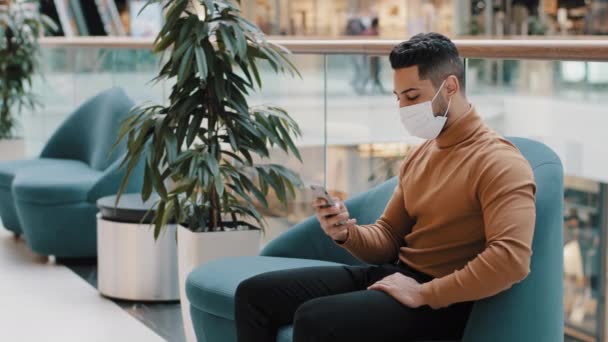 Hombre joven en máscara médica se sienta en el interior escribiendo mensaje en la red social en el teléfono inteligente envía correo electrónico a través del teléfono mediante la aplicación móvil para ordenar en la tienda de Internet de navegación web en línea citas sitio — Vídeo de stock