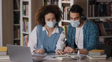 Tıp maskeli iki öğrenci üniversite kütüphanesinde masada oturup kulaklıklı hocaları dinliyorlar. Laptop çalışmasında, video konferans üzerinden, defter konferanslarında öğrendikleri bilgileri not ediyorlar.