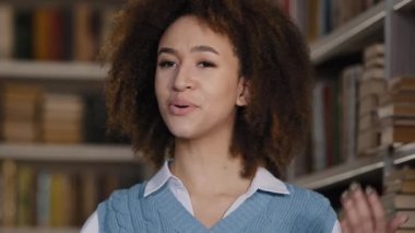 Web kamerası görüntüsü genç Afro-Amerikan kadın blogger öğrenci devlet üniversitesi kütüphanesinde kameraya bakarak konuşuyor video konferansı yaparak üniversite eğitim mesafesi sohbeti hakkında bilgi veriyor