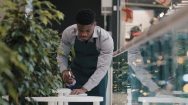 Genç Afro-Amerikalı garson garson garsonlar apronlu temizleme masası dezenfektanla temizler. Restoranı açmadan önce yarım gün çalışan ziyaretçiler için kafe hazırlar.