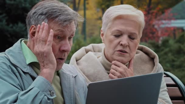 Shocked old married couple looking at laptop screen sitting in autumn park upset elderly man and woman reading bad news worried spouses aged feel shock stress discuss problem grandfather holding head — Stock Video