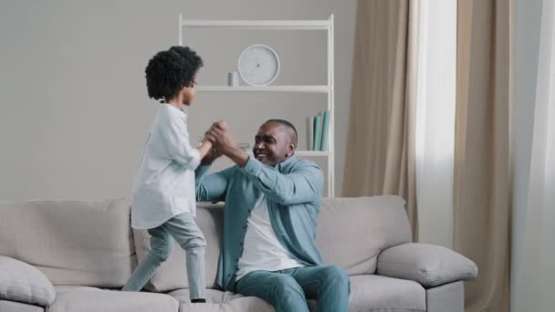 Afrykański amerykański ojciec z małą córeczką spędzają wolny czas razem bawiąc się aktywnie tańcząc do muzyki opiekuńczy tata siedzi na kanapie trzymając dziecko dziewczyna przez ręce dziecko radośnie skacząc — Wideo stockowe