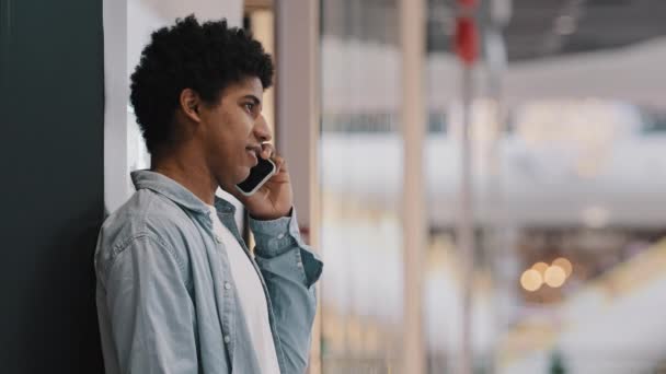 Vista lateral afro-americano millennial 20s homem de negócios cara masculino estudante de pé perto da parede no espaço público falando no telefone celular atender chamada conversando com amigos rindo conversa wi-fi gratuito — Vídeo de Stock