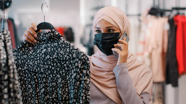 头戴医疗面罩的年轻阿拉伯女人在服装店里选择服装。打电话咨询如何选择新衣服。 — 图库照片
