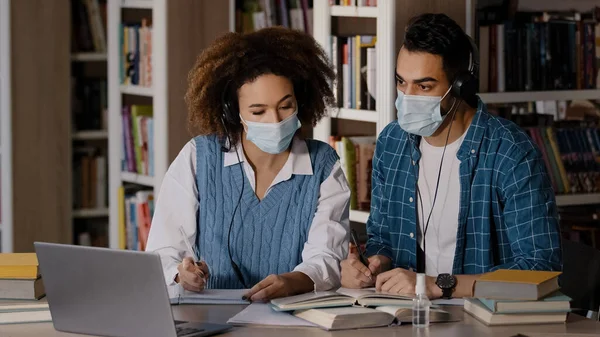 Два студента в медицинских масках сидят за столом в университетской библиотеке, слушают учителя в наушниках на ноутбуке, записывают информацию в ноутбуке — стоковое фото