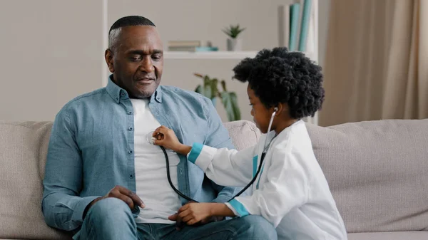 Little african american girl in medical gown plays sitting on couch kid doctor listens to father stethoscope pretends nurse having fun loving daughter forbids dad to smoke indicates get rid bad habit — Stock Photo, Image