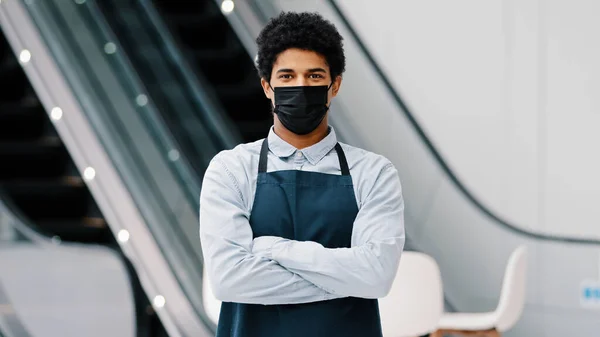 Африканский американец парень ресторан кафе мужчина в медицинской маске смотрит в камеру позируя скрещивая руки официант в фартуке стоя на рабочем пространстве ковид пандемия карантина. Малый бизнес — стоковое фото