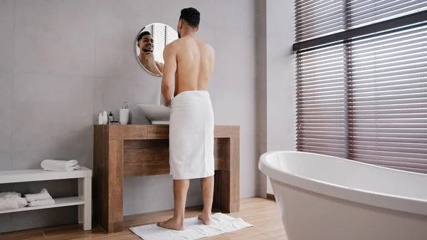 Полноценный вид обнаженного мускулистого спортсмена-индийца с игрушкой на хипстере, смотрящего в зеркало в ванне, наслаждается прекрасным парнем, трогающим бороду в ванной, делающим утреннюю косметологическую процедуру — стоковое фото