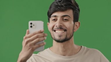 Arap genç adam stüdyoda yeşil arkaplanda video çekiyor konferans uygulamasını kullanarak telefonda arkadaşlarıyla keyifli bir şekilde konuşuyor iyi haberleri paylaşıyor heyecanlı adam el sallıyor canlı yayına başlıyor