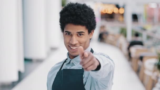 Amistoso afroamericano joven hombre camarero vendedor restaurante cafetería cocina bar trabajador en delantal mirando la cámara mostrando gesto ella usted viene aquí enfoque bienvenida invitando a los clientes promociones — Vídeo de stock