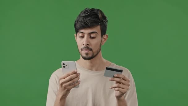 スタジオで緑の背景に幸せな笑顔の男アラブ人男性は、携帯電話やクレジットカードを使用してインターネット注文を支払うアプリで取引のための番号を入力します若い男性は、モバイルバンキングアプリケーションを使用してリモートショッピング — ストック動画