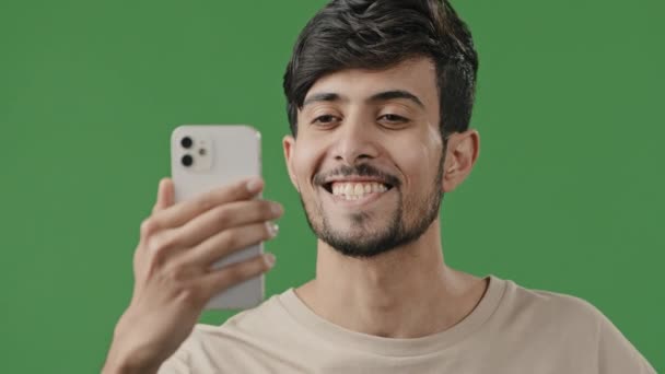 Árabe joven sonriente chico haciendo videollamada en fondo verde en el estudio utilizando la aplicación de conferencia en el teléfono alegremente hablando con un amigo en línea comparte buenas noticias hombre emocionado mano de onda a partir de corriente en vivo — Vídeos de Stock