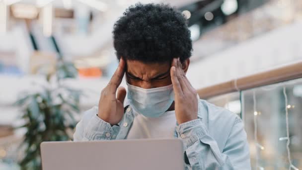 Afrykański amerykański biznesmen facet student noszenie maski medycznej pracy z laptopem w korporacyjnej przestrzeni biurowej trzymając głowę uczucie bólu głowy cierpi ciśnienie migrena objawy koronawirusowe przepracowanie — Wideo stockowe