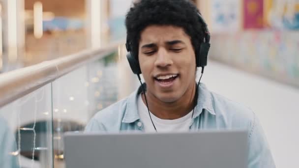 African american 20s guy student business male freelancer sitting in cafe indoors with laptop wears headphones listening to music online website enjoying audio sound in computer app singing song break — Stock Video