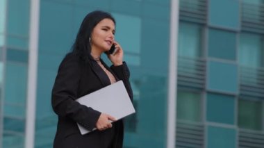 Başarılı genç iş kadını caddelerde yürürken şehrin arka planında dizüstü bilgisayarı, kendine güveni olan İspanyol kız müdürü cep telefonuyla konuşuyor ve cep telefonuyla konuşuyor.