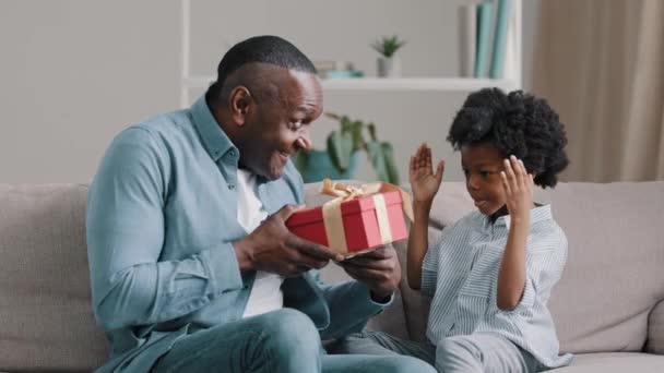 Trochę Afryki słodkie dziewczyna siedzi na kanapie obejmuje oczy z rąk kochający ojciec dając prezent urodzinowy dla córki daje czerwony prezent pudełko dziecko zadowolony z nieoczekiwanej niespodzianki radosny tata przytula dziecko — Wideo stockowe