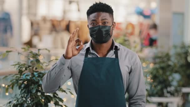 医療マスクに立つエプロンカフェウェイターで若いアフリカ系アメリカ人男性レストラン労働者バリスタ営業マンは、隔離中にokジェスチャー承認サイン小さなビジネスプロモーションを示すカメラを見て — ストック動画