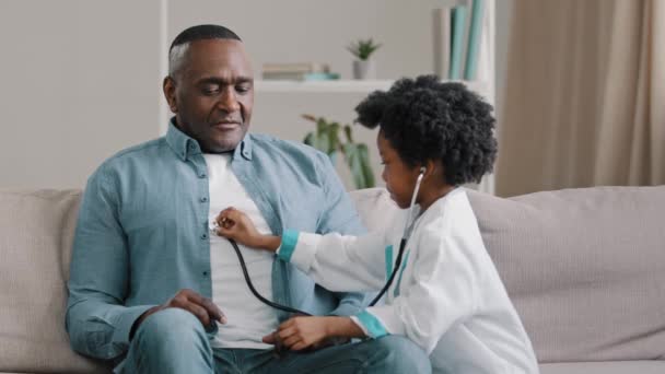 Mała afrykańska dziewczyna w medycznej sukni gra na kanapie dziecko lekarz słucha ojca stetoskop udaje pielęgniarka mając zabawy kochający córka zabrania tacie palić wskazuje pozbyć się złego nawyku — Wideo stockowe