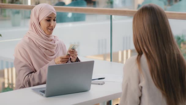 Unga vänliga leende arabiska kvinna chef sitter på kontoret vid skrivbordet kommunicerar med kunden förklarar villkor kontrakt ger pengar oigenkännliga kaukasiska flicka tar ut kontanter från bankkonto tar lån — Stockvideo