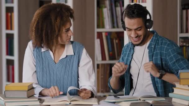 Estudiantes en la biblioteca preparándose para el examen haciendo deberes divertido joven escuchando música en auriculares bailando distraído de las lecciones interfiere estudiar indignado compañero de clase muestra gesto de silencio — Vídeos de Stock