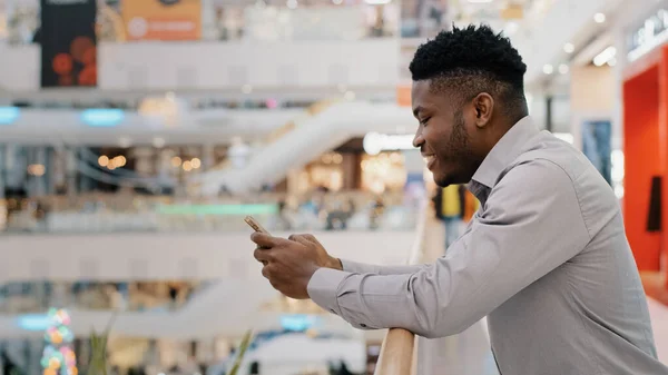 Молодой африканский американец, стоящий в торговом центре, проводит свободное время в соцсетях Интернет на смартфоне с помощью виртуального сервиса приятное общение на сайте улыбается, наслаждаясь дистанционным чатом — стоковое фото
