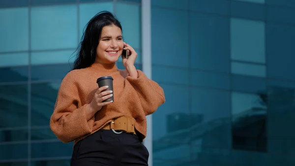 Heureuse femme hispanique debout à l'extérieur en buvant du café de tasse en papier répondant à un appel amical souriant ayant conversation agréable sur téléphone mobile communiquer à distance avec un ami à l'aide d'un smartphone — Photo