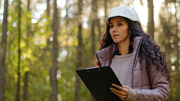 Milenial mujer técnica ecologista mirando hacia arriba en las copas de los árboles, Joven mujer india en hardhat con portapapeles tomando medidas de comprobación de los árboles. Ingeniero forestal en el parque. Supervisar el santuario de vida silvestre —  Fotos de Stock