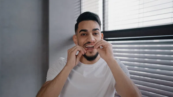 Portrait millennial handsome bearded arab indian male guy in white shirt brushing teeth using dental floss morning hygiene ritual oral cleaning toothy at home in bathroom health care dentist concept