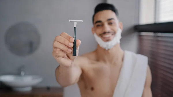 Funny hispanic indian arab naked man 30s bearded guy standing in bathroom at home with white soapy foam on beard shaving gel smiling showing razor looking at camera getting ready to shave facial care — стоковое фото