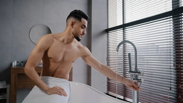 Millennial σέξι γυμνός μυώδης ινδός Άραβας άντρας με γενειάδα τυλιγμένη πετσέτα γύρω από τους γοφούς κάθεται στην μπανιέρα στο μπάνιο στο σπίτι γυρίζοντας βρύση δοκιμάζοντας ζεστό κρύο νερό με το χέρι ετοιμάζεται για μπάνιο — Φωτογραφία Αρχείου