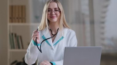 Kafkasyalı kadın doktor, kardiyolog, hemşire, hemşire, gözlük ve cüppe uzmanı. Kameraya bakıp gülümsüyor. Hastane kliniğinde dizüstü bilgisayarla steteskopla oturuyor.