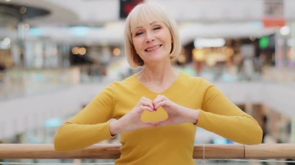 Madura mujer caucásica feliz de pie en lugar público posando sinceramente sonriendo mirando a la cámara haciendo forma de corazón con los dedos signo generosidad y amor gesto de misericordia bondad símbolo amistad — Vídeo de stock