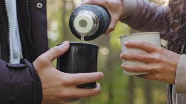 Ormandaki tanınmayan iki kişi termoslu adamdan sıcak çay dolduruyor kızla birlikte doğada dinleniyor sıcak içeceklerle ısınırken eğlenceli zaman geçiriyor genç çiftler hafta sonlarını kamp yaparak geçiriyor.