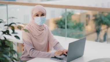 Tıbbi maskeli genç Arap kadın, iş yerinde oturmuş dizüstü bilgisayarda yazarak yeni bilgisayar uygulama yazılımını kontrol ediyor ve iyi bir geri bildirim bırakarak onay işareti işareti işareti işareti yapıyor.